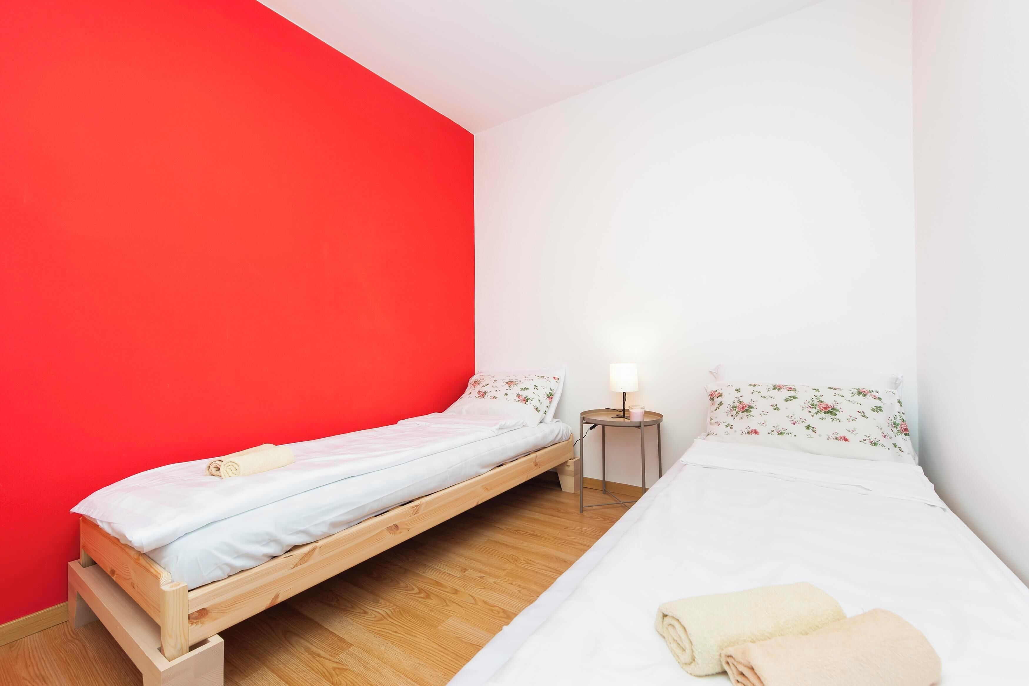 A small room with two beds, one with a wooden frame and the other with a metal frame. There is a small table with a lamp between the beds.