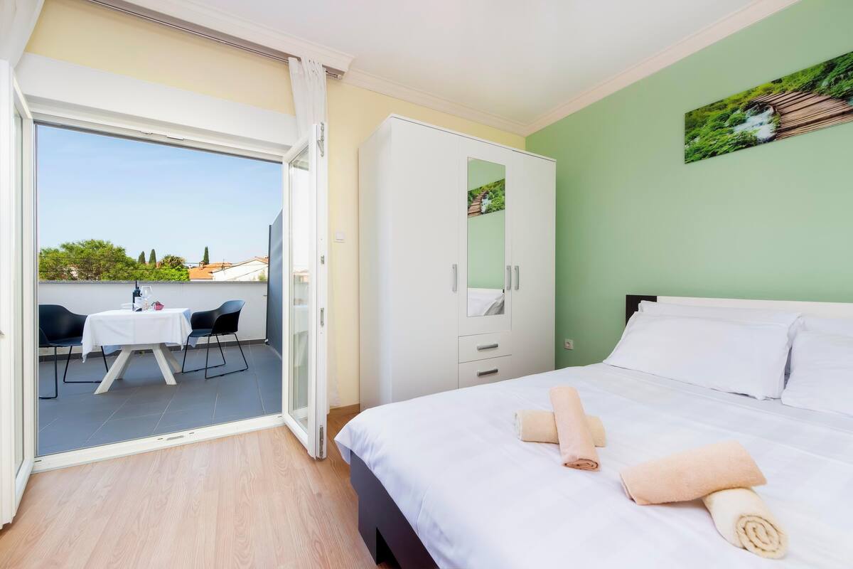 A hotel room with a bed, a wardrobe, a balcony with a table and chairs, and a view of buildings.