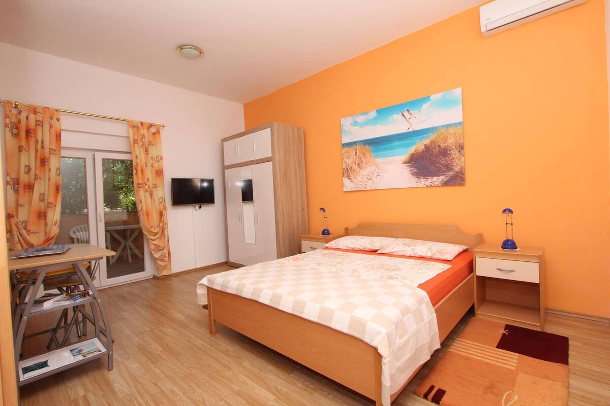 A bedroom with a bed, a desk, a chair, a nightstand, and a wall-mounted TV.