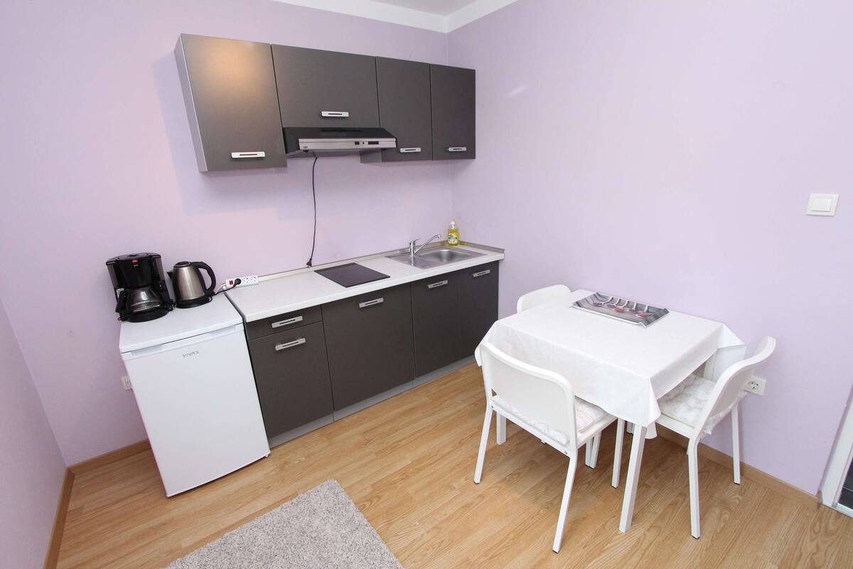 A compact kitchen with a white refrigerator, a coffee maker, and a sink. There is a small table with two chairs and a magazine on it.