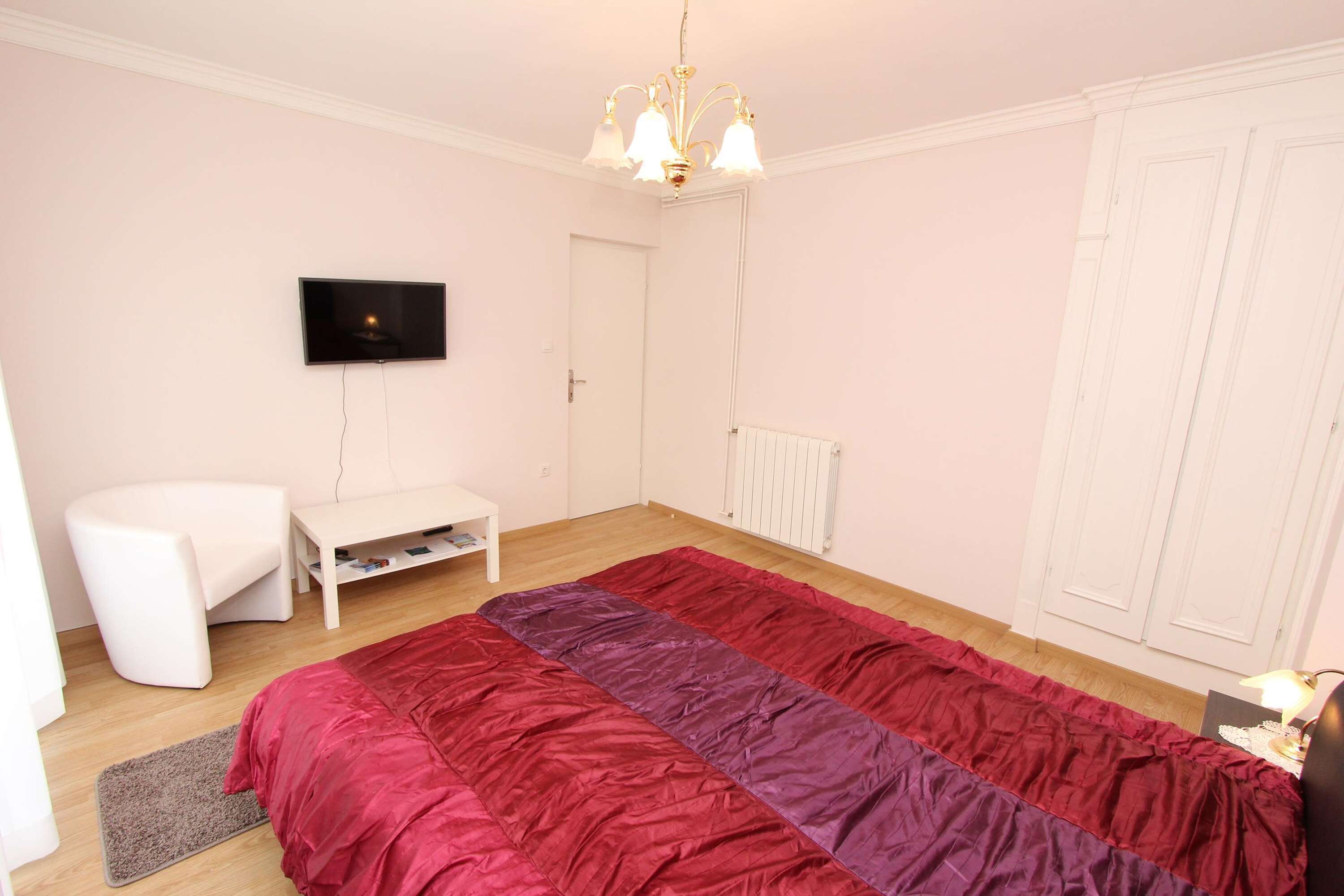 A bedroom with a bed, a TV, a chair, a small table, and a wardrobe.