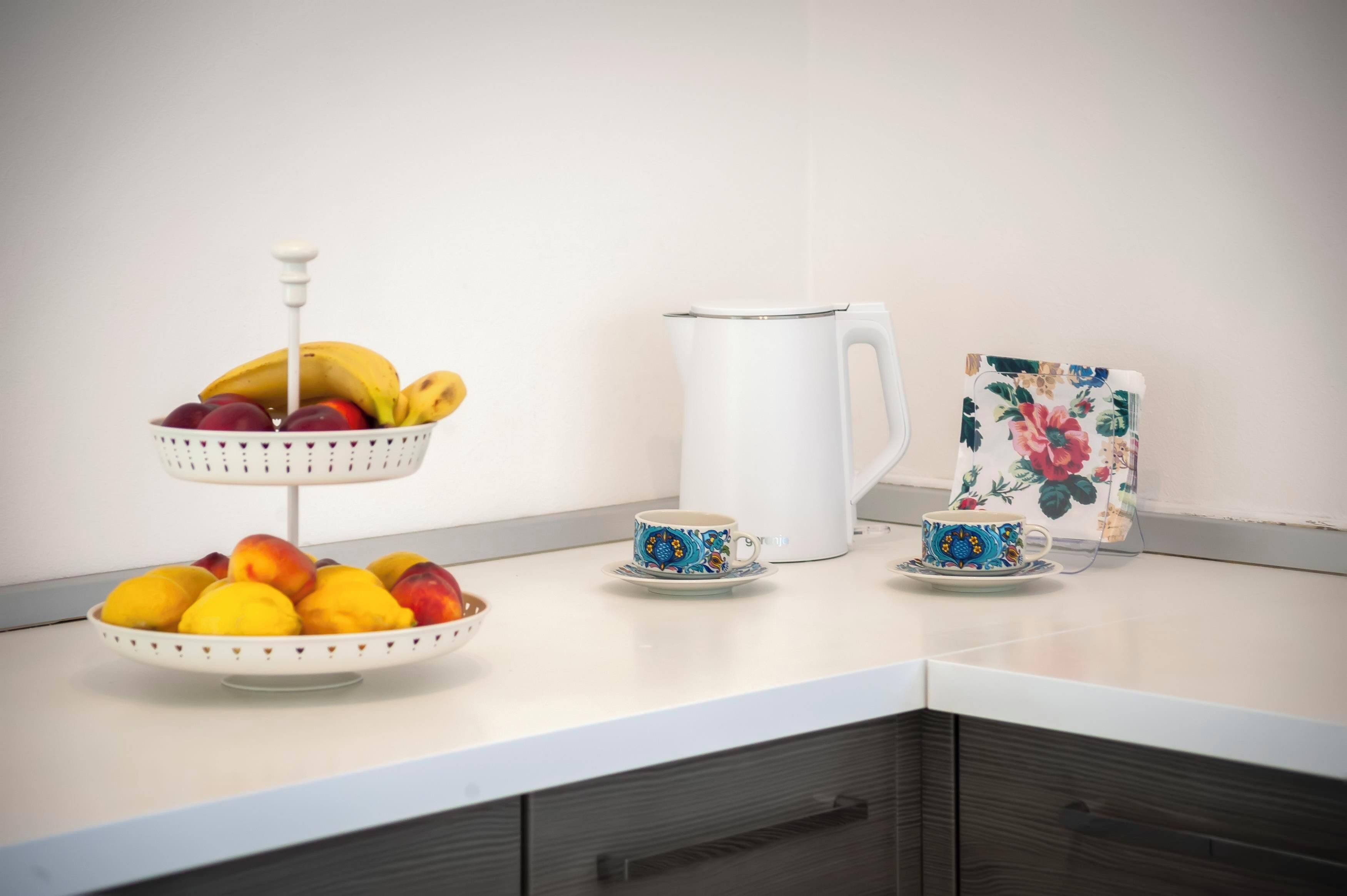 Eine Küchenarbeitsplatte mit Obstschale, einem weißen Wasserkocher, einer floral verzierten Tasse und Untertasse.