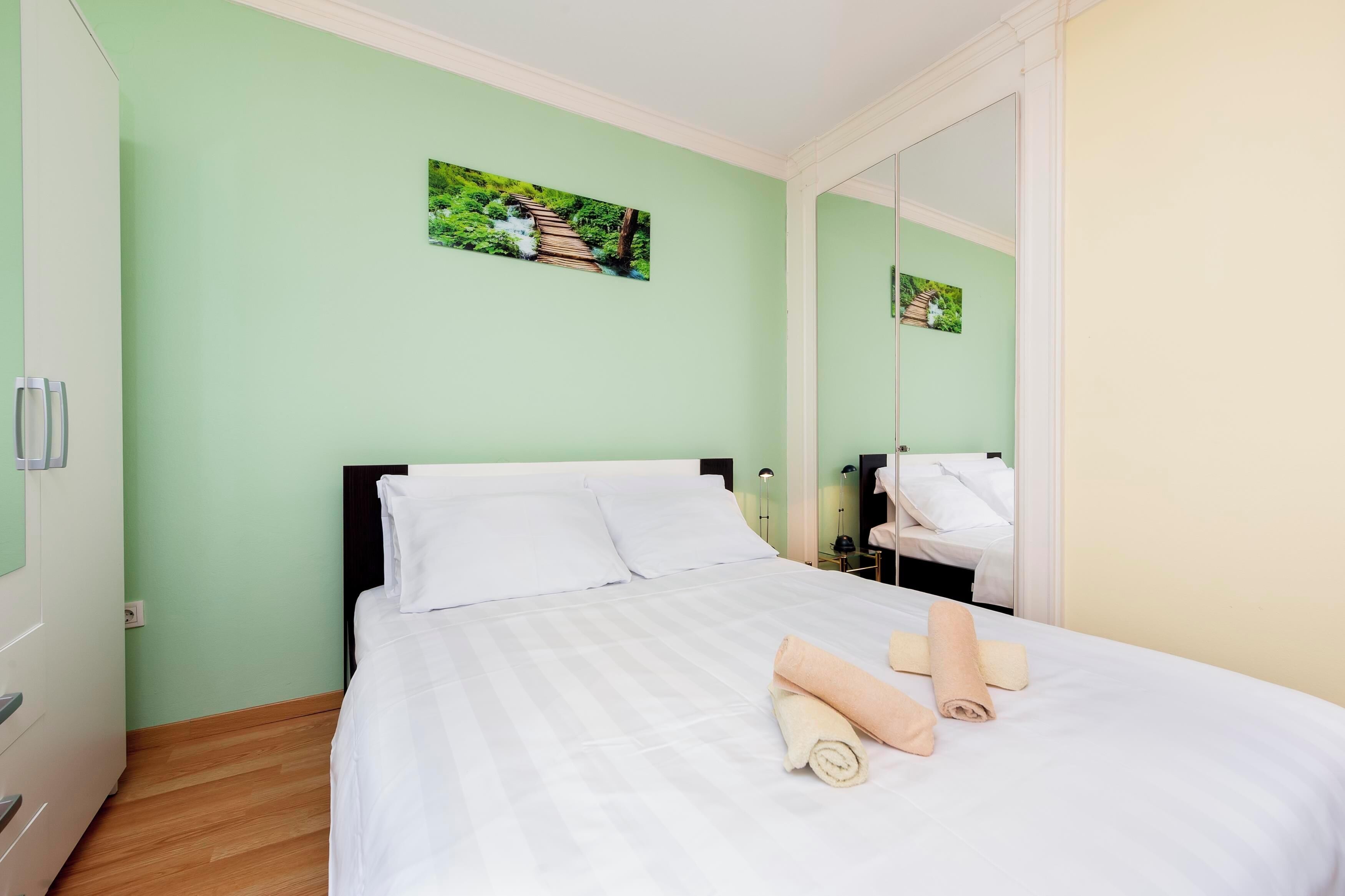 Een netjes opgemaakt bed met witte lakens en twee opgerolde handdoeken, een groene muur met een natuurfoto, en een spiegel die een deel van de kamer weerspiegelt.