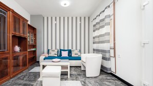 A modern living room with a sofa, a coffee table, and a white armchair. There is a wooden cabinet with a pink mug on it. The room has striped wallpaper and a striped curtain. - Apartments Sena (Rovinj)