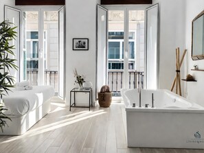 A modern hotel room with a large bathtub, a sofa, a small table with a vase, and a picture on the wall.