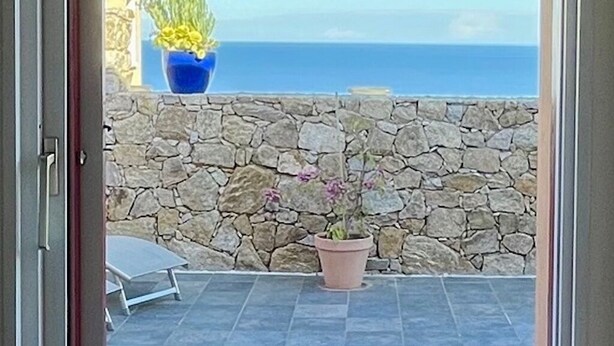 A balcony with a stone wall and potted plants, overlooking a clear blue sky and sea.