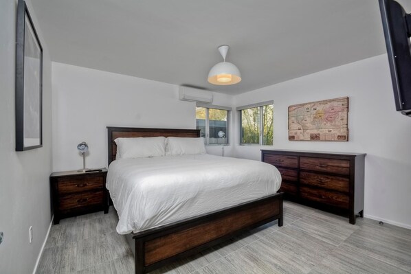 A bedroom with a large bed, a wooden dresser, a desk with a lamp, and a map on the wall. - Wind (1 bedroom / 1 bath) (Carlsbad)