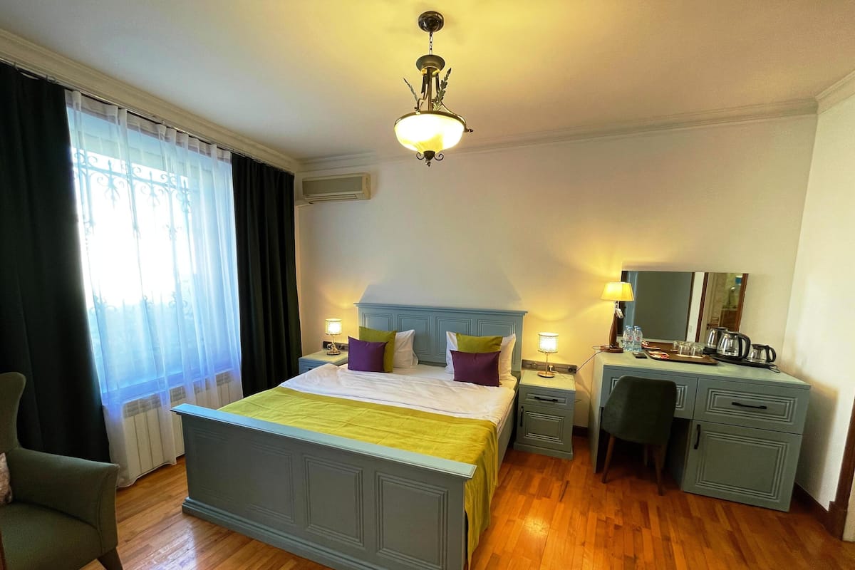 A hotel room with a large bed, a desk, a chair, a desk lamp, a bedside table, and a window with curtains.
