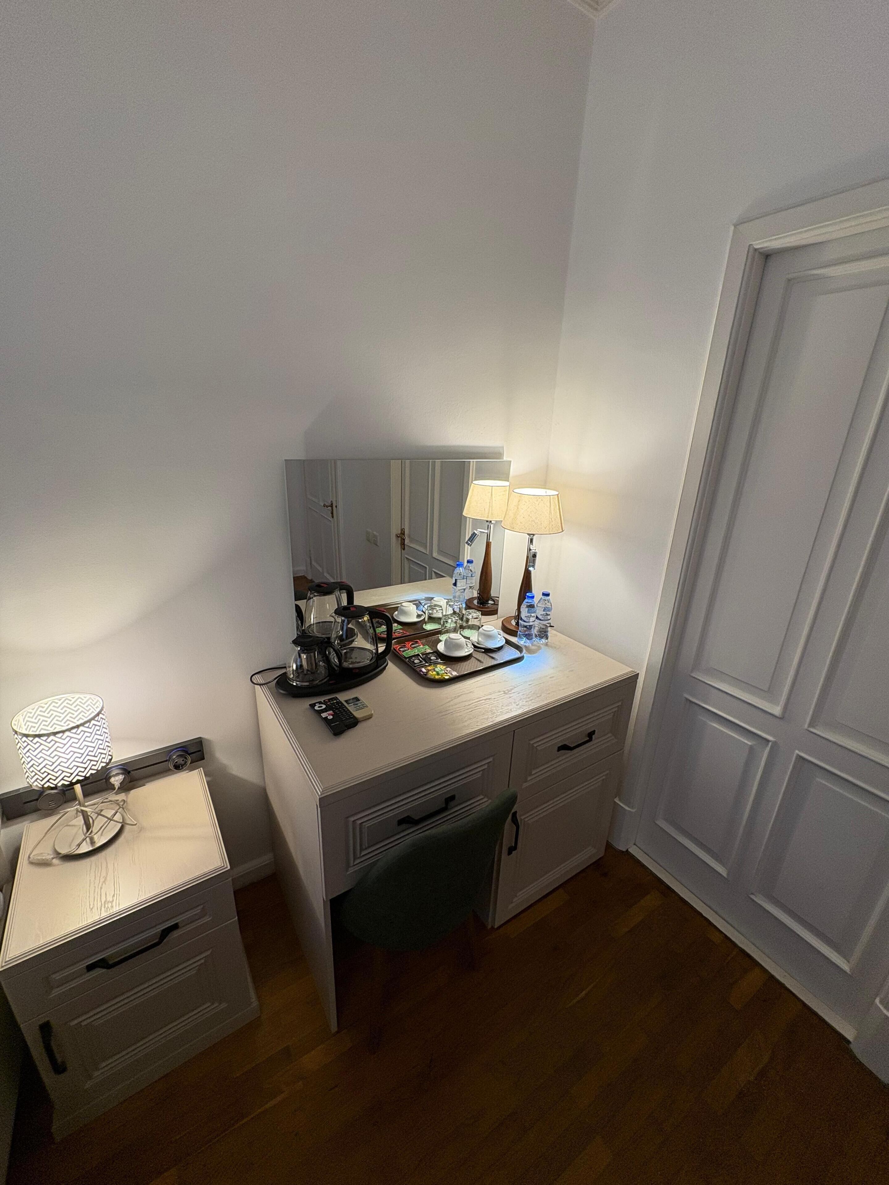 A small, neatly organized bedroom with a wooden floor, a bedside table with a lamp, a mirror, and a dresser with drawers.