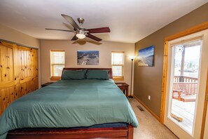 A bedroom with a bed, wooden wardrobe, ceiling fan, two windows with blinds, and a view of a deck through a glass door.