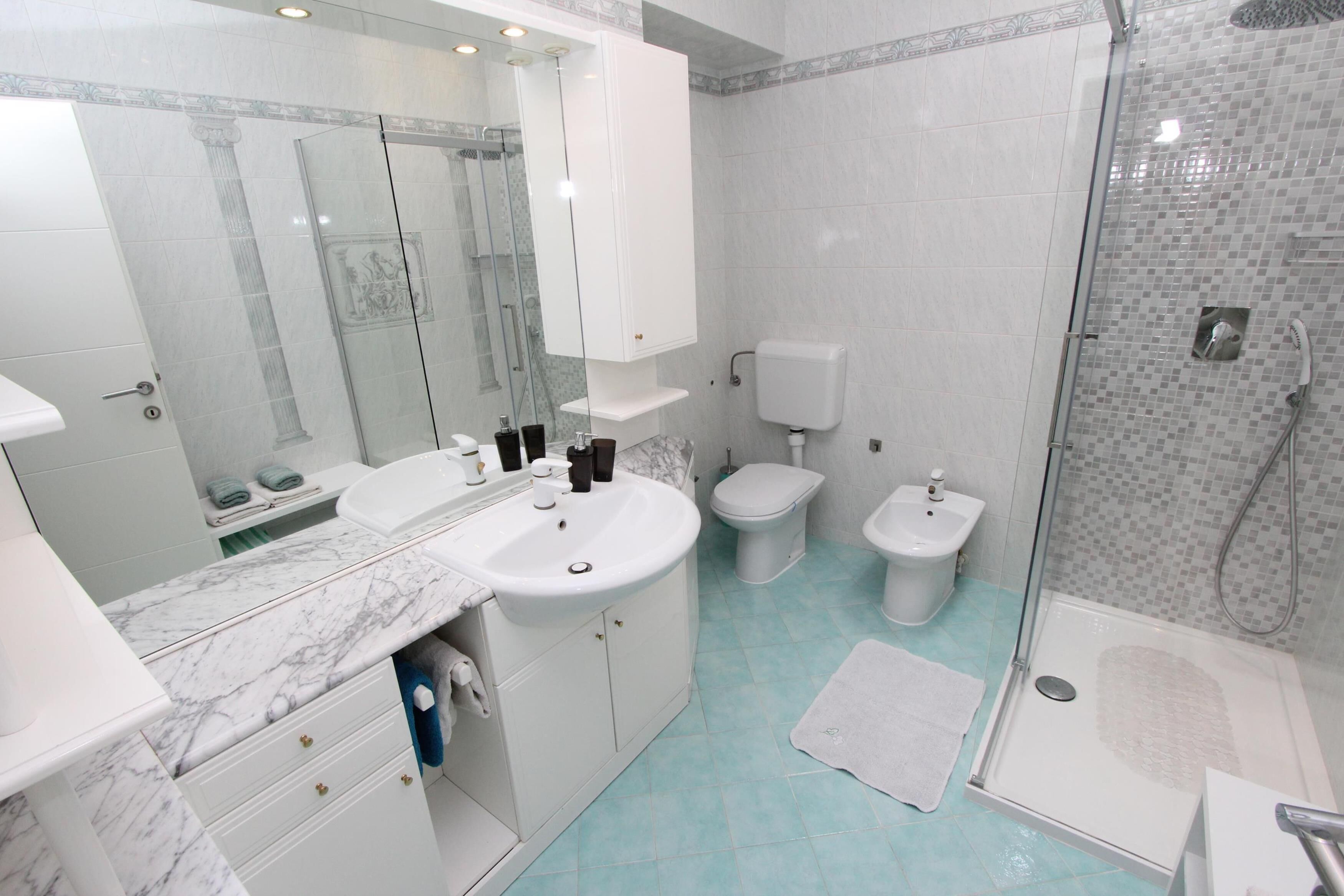 Un bagno moderno con lavandino in marmo, due servizi igienici, cabina doccia in vetro e mobile bagno bianco con cassetti.