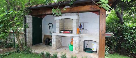 A rustic outdoor kitchen with a stone fireplace, a wooden pergola, and a green watering can.