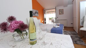 Hotelkamer met een eettafel gedekt met een fles wijn en glazen, een vaas met roze bloemen en een bed met een blauwe deken.