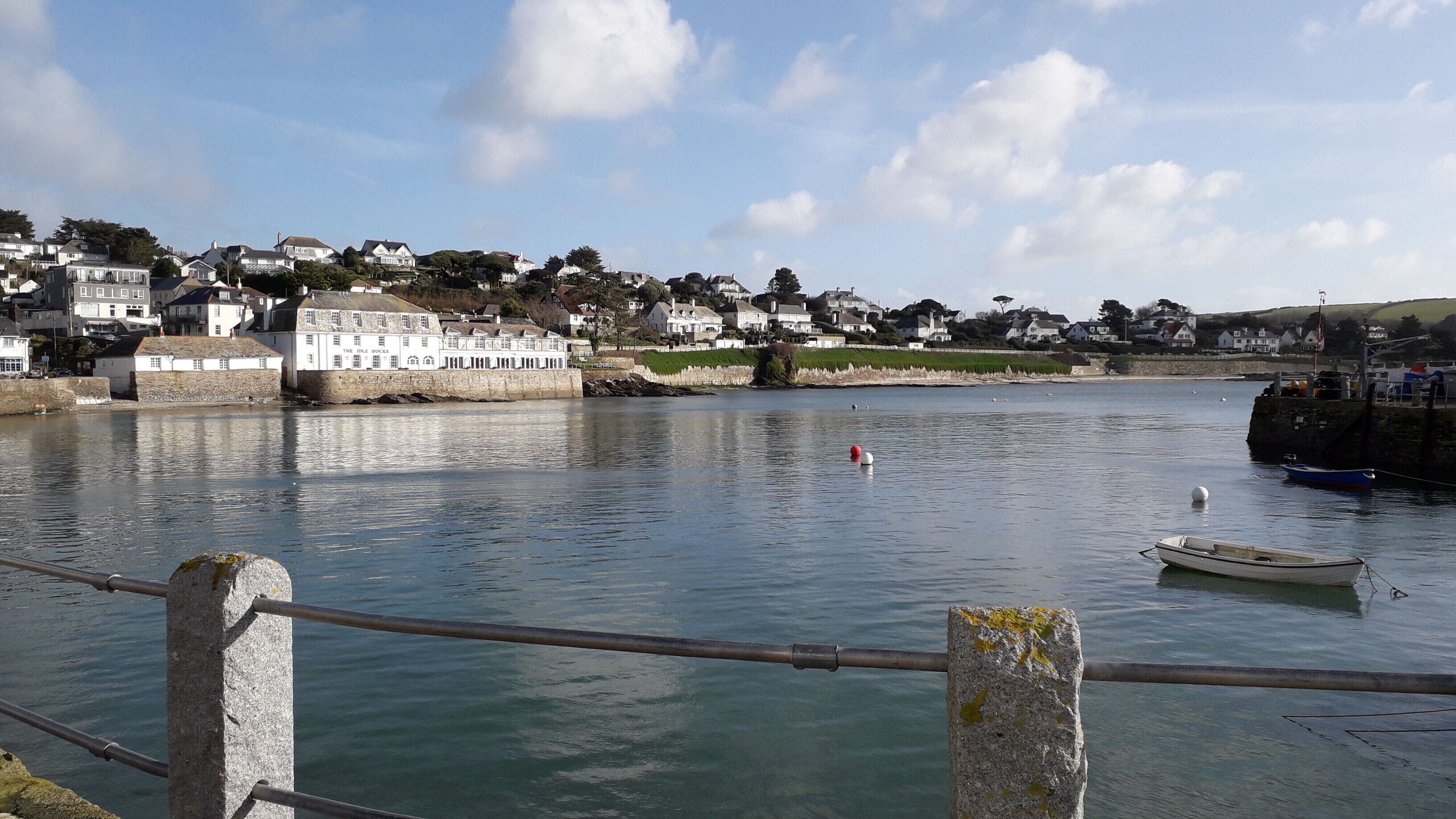 A waterfront view with a stone railing, a small boat, and a row of houses in the background.