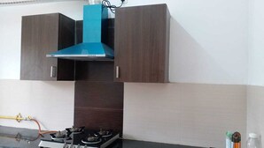 A kitchen with a gas stove, a blue range hood, and wooden cabinets. - LAZY BEAR VILLA  IN  LAP OF NATURE (GREATOR NOIDA)
