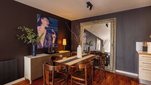Un comedor con mesa de madera, sillas, un florero con ramas, un cuadro y un espejo.