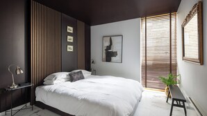 Un dormitorio moderno con una cama grande, una mesita de noche con lámpara, un cuadro enmarcado en la pared y una ventana con persianas.