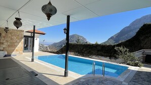 A pool area with stone flooring, a covered patio, hanging lanterns, and a mountain view. - Esiyok Villa in Antalya with heated pool (Hisarcandir Mahallesi Konyaalti)