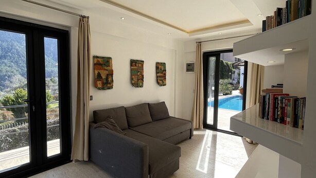 A modern living room with a grey sofa, a bookshelf, and a view of a pool through a glass door. - Esiyok Villa in Antalya with heated pool (Hisarcandir Mahallesi Konyaalti)