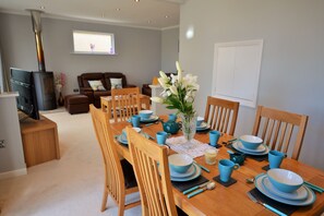 A dining room with a wooden table set for four, surrounded by chairs. A fireplace is visible in the background, and a window with blinds is on the wall.