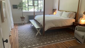 A bedroom with a four-poster bed, a bedside table with a lamp, a bench, a window with a view of trees, and a potted plant. - 1940s Brooklyn Cottage Townhouse (Brooklyn)