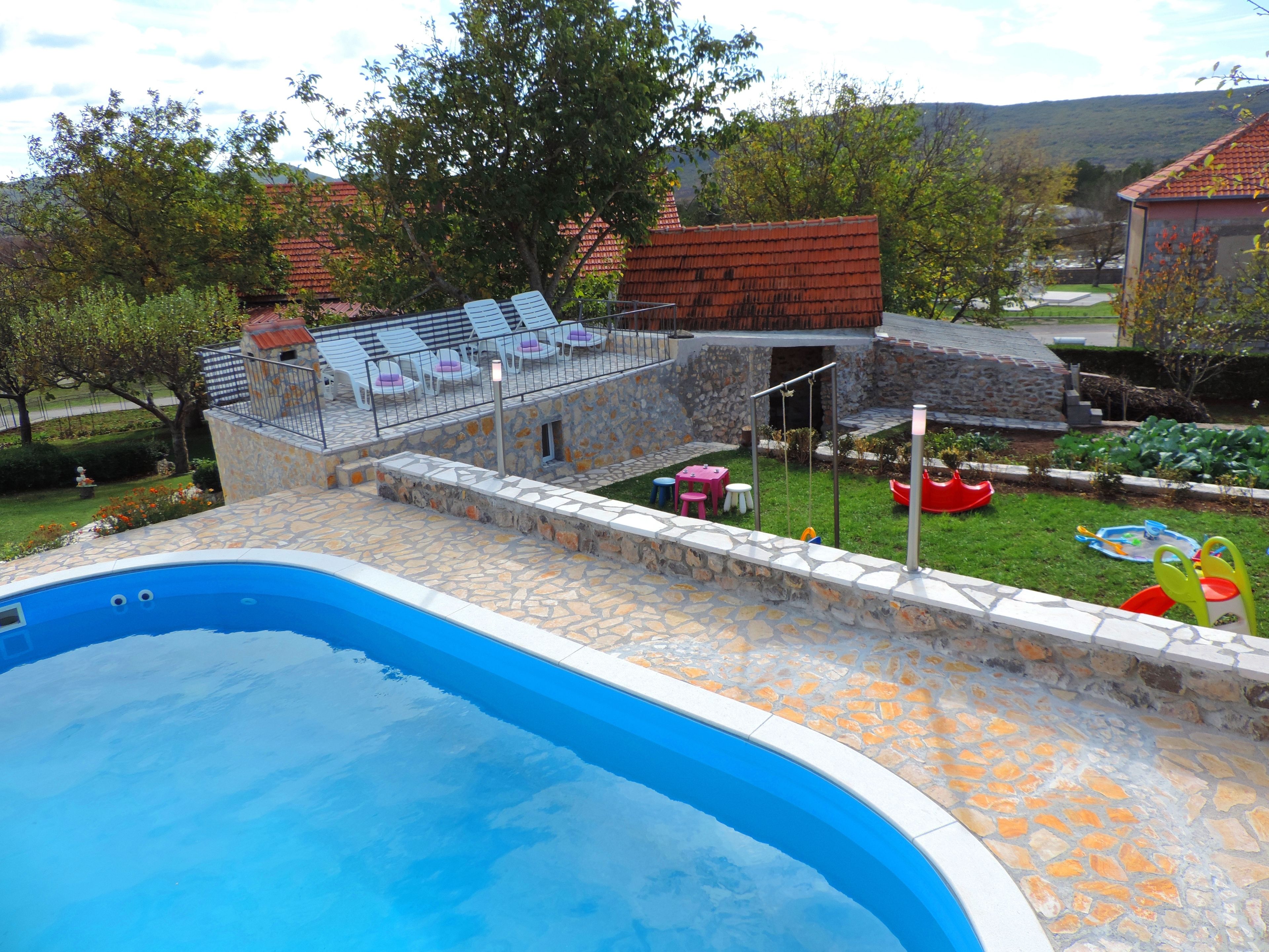A swimming pool with a stone deck, surrounded by a garden with a play area and lounge chairs.