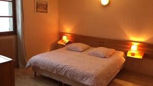 A bedroom with a wooden headboard, two bedside lamps, a floral bedspread, and a framed picture on the wall. - Le Grand Joly (Sainte-Agnès)