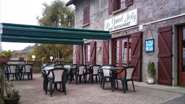 An outdoor seating area with tables and chairs, a covered patio, and a building with a sign reading "Le Grand Joly BAR RESTAURANT". - Le Grand Joly (Sainte-Agnès)