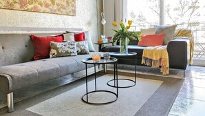 Een moderne woonkamer met een grijze bank, rode en witte kussens, een klein rond tafeltje met een bloemenvaas, en uitzicht op het buitenleven door grote ramen.