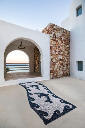 Ein zeitgenössischer architektonischer Raum mit einer Steinsäule, einem rundbogenförmigen Durchgang, der auf einen Balkon mit Meerblick führt, und einem gemusterten Teppich auf dem Boden.