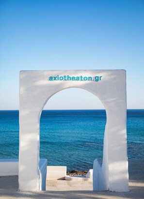 Ein weißes Tor mit dem Schriftzug "axiotheaton.gr" vor einem klaren, blauen Himmel und dem Meer.