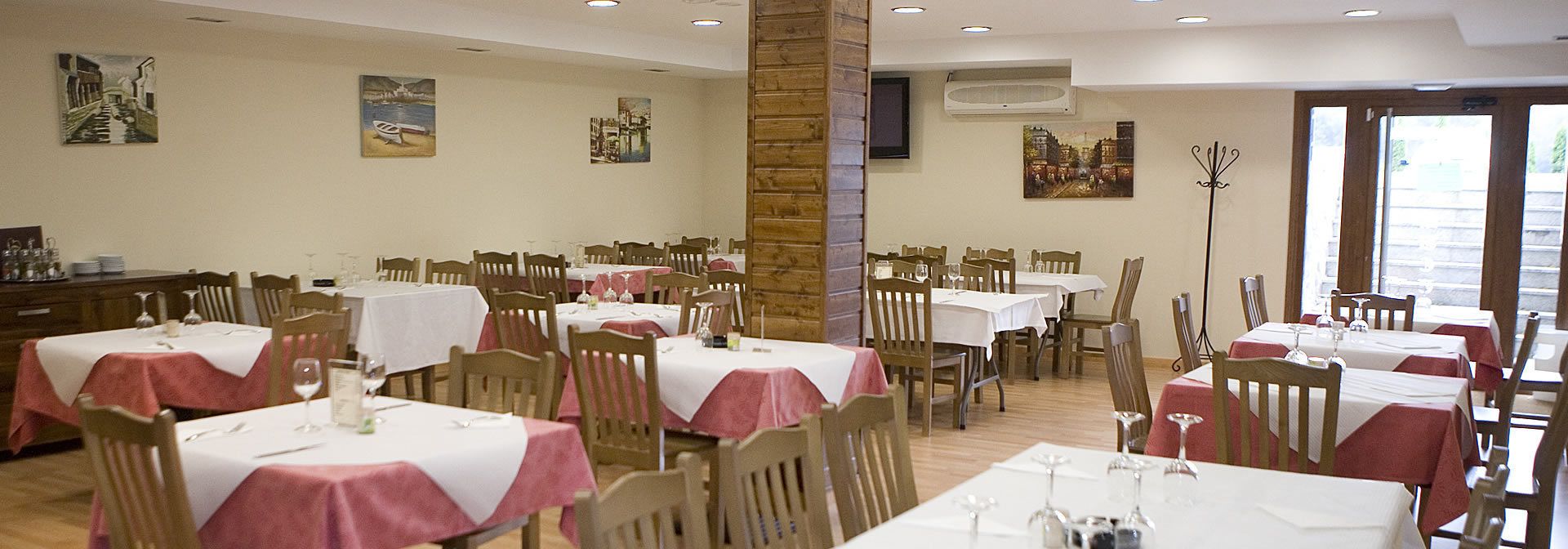 A dining area with multiple tables, each covered with a white tablecloth and red napkins. There are wooden chairs around the tables, and the walls are adorned with paintings.