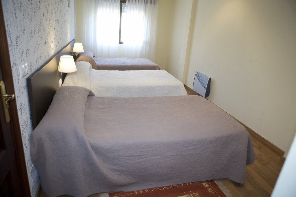 A hotel room with two beds, a wooden floor, a window with sheer curtains, and a nightstand with a lamp.