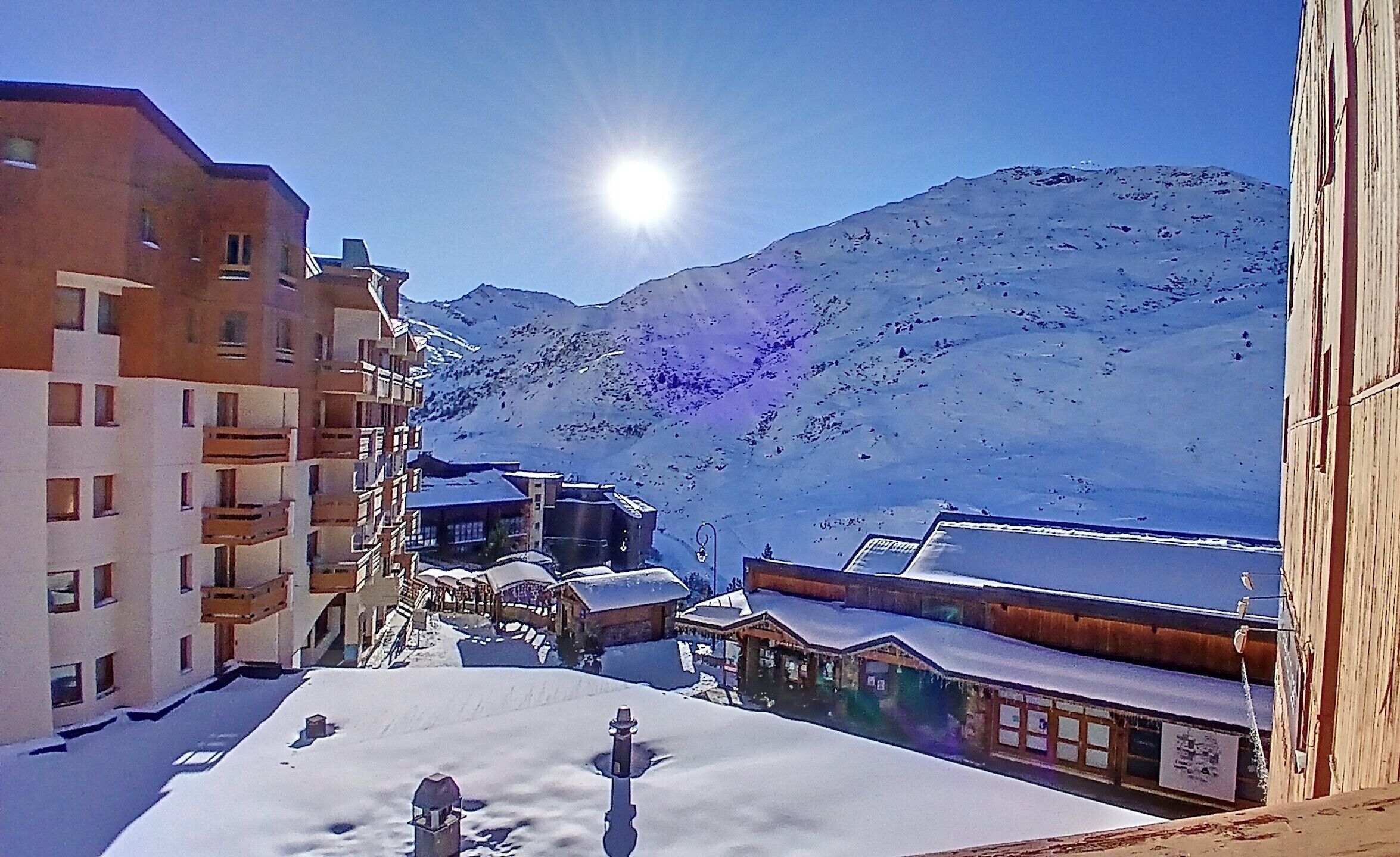 Una strada innevata con edifici, una montagna sullo sfondo e il sole che splende.
