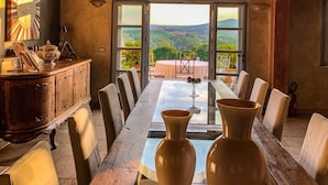 A dining area with a long wooden table, chairs, and two large vases. There is a chandelier above the table and a view of the outdoors through large windows. - Country House Villa Rey (Panicale)