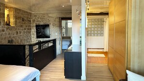A modern bedroom with a stone wall, a wooden floor, a bed, a desk, and a bathroom with a shower. - Forcalquier Architecture Village House Center (Forcalquier)