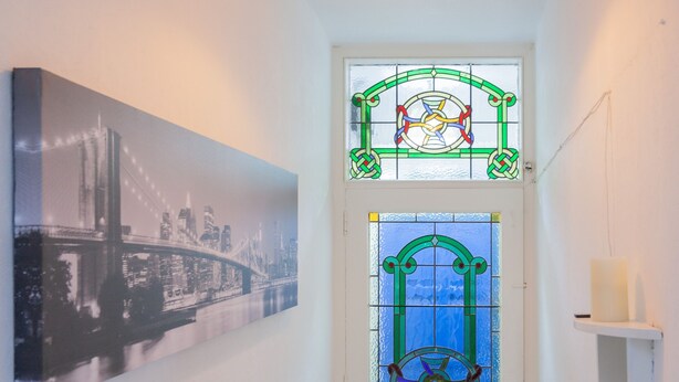 A hallway with a door featuring a stained glass design, a framed cityscape photograph on the wall, and a white shelf with a candle. - Windsor House - A Home from home (Derry)