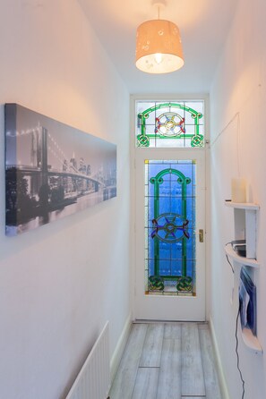 A hallway with a door featuring a stained glass design, a framed cityscape photograph on the wall, and a white shelf with a candle. - Windsor House - A Home from home (Derry)