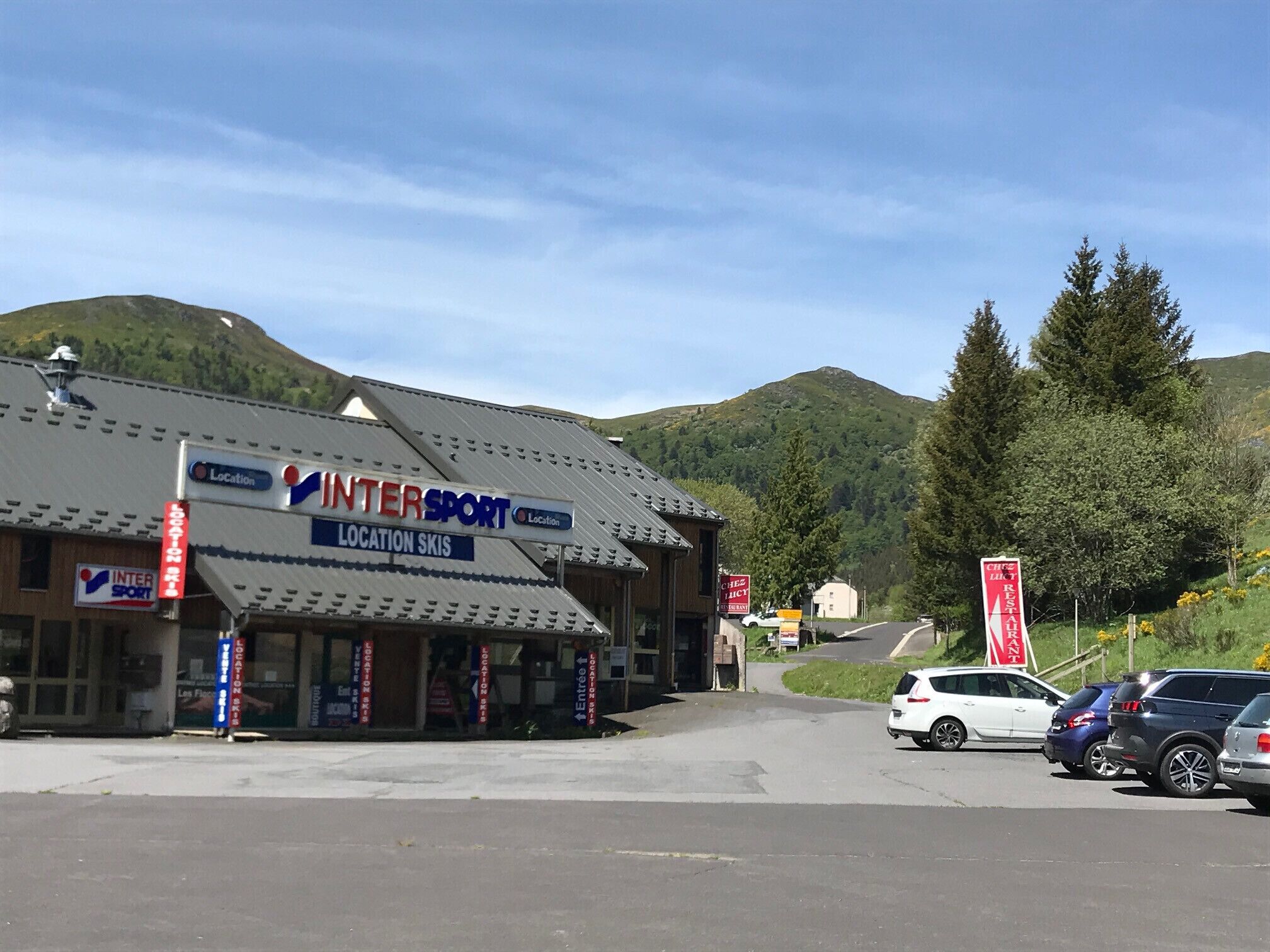Un bâtiment avec une enseigne indiquant « INTERSPORT LOCATION DE SKIS » et un parking avec des voitures.