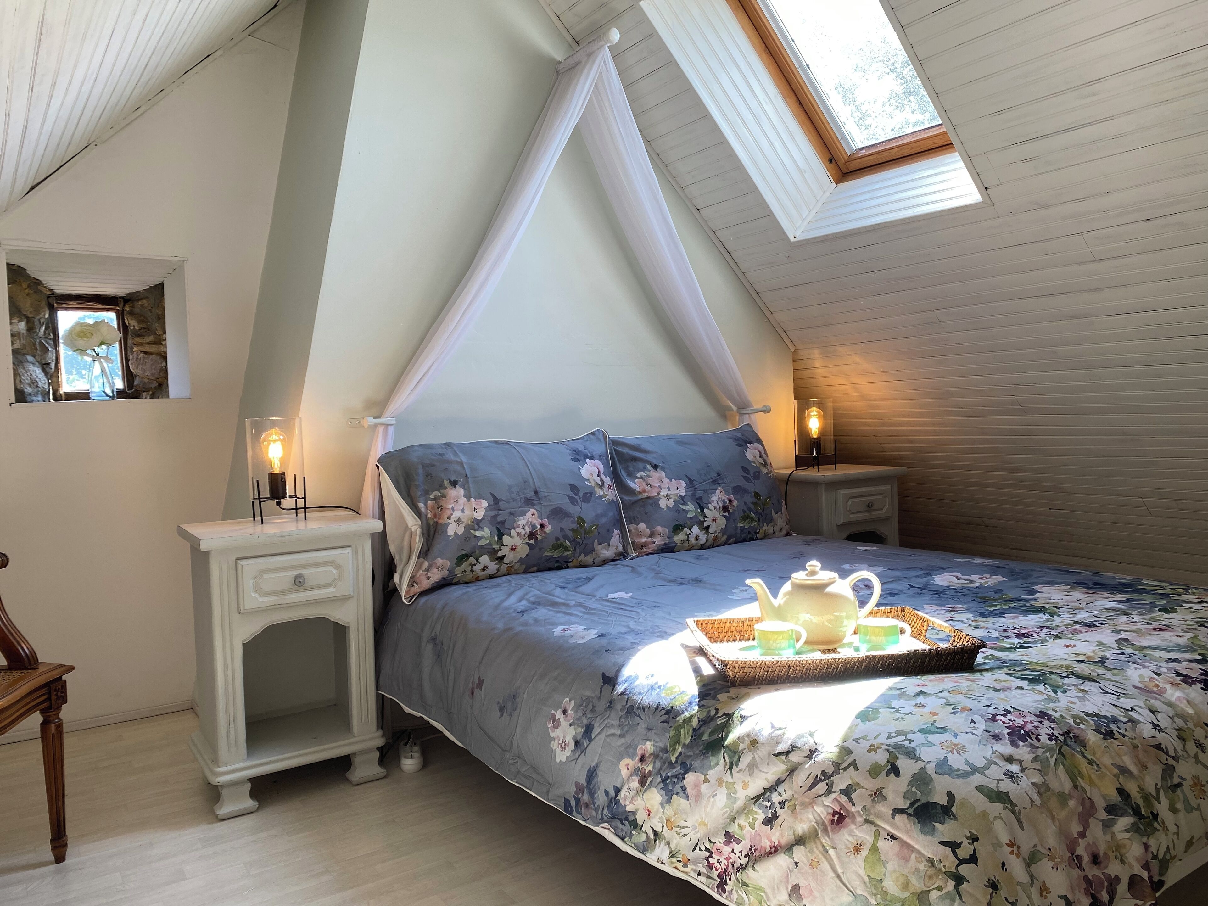 Een slaapkamer met een schuine balkenlaag, een bed met bloemenbeddengoed, nachtlampjes, een klein tafeltje met een theepot, en een raam met uitzicht.