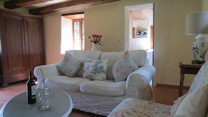 A living room with a white sofa, floral cushions, a small table with a wine bottle and glass, and a wooden cabinet. - Private detached stone house overlooking a lake (Sazeray)