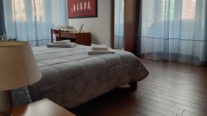Habitación de hotel con cama, escritorio, silla, lámpara, un cuadro en la pared y una ventana con cortinas.