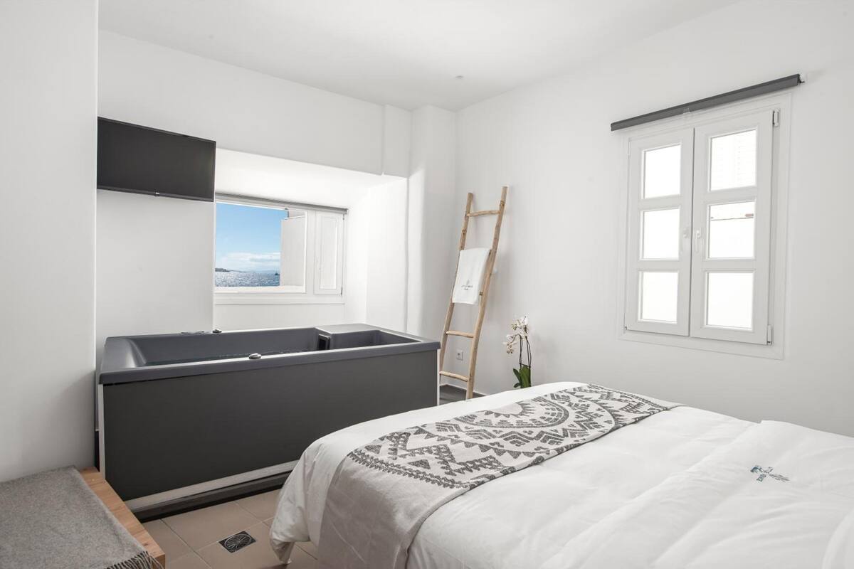 Une chambre moderne avec un grand lit, une baignoire intégrée, une échelle appuyée contre le mur et une fenêtre donnant sur la ville.