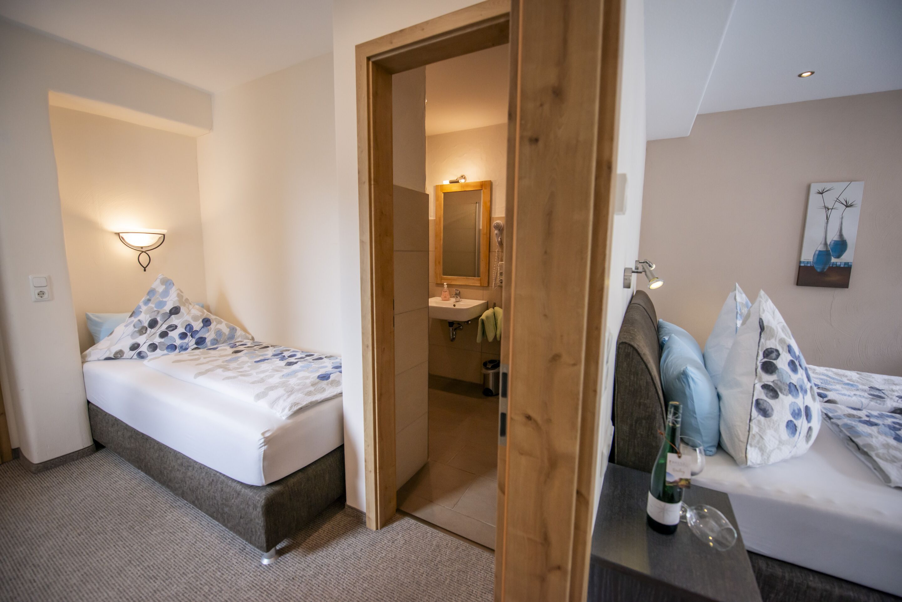 A bedroom with a bed, a wooden door, a bathroom visible through the door, and a small table with a bottle and glass.