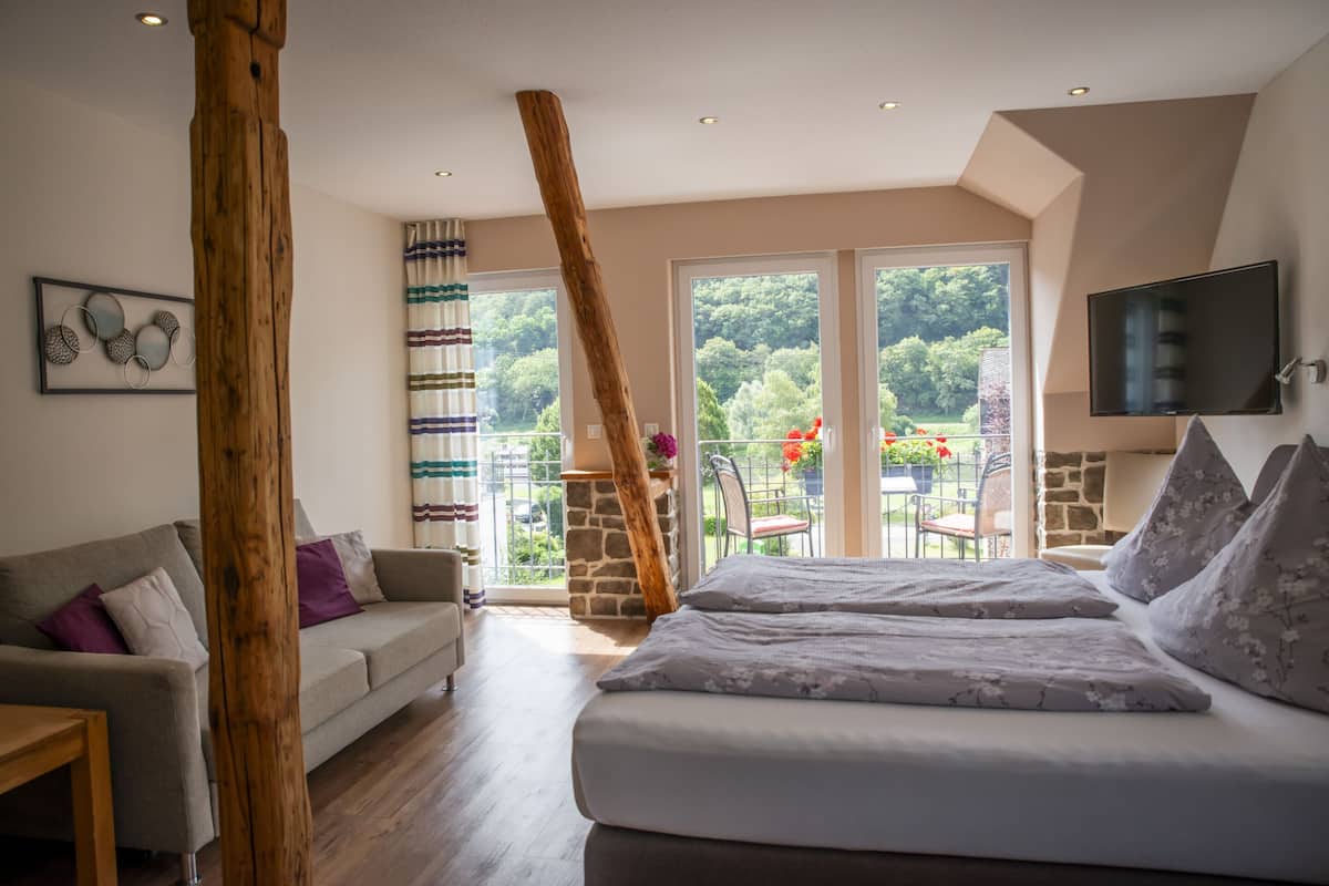 A bedroom with a bed, a wooden beam, a stone fireplace, a TV, and a balcony with a view of trees.