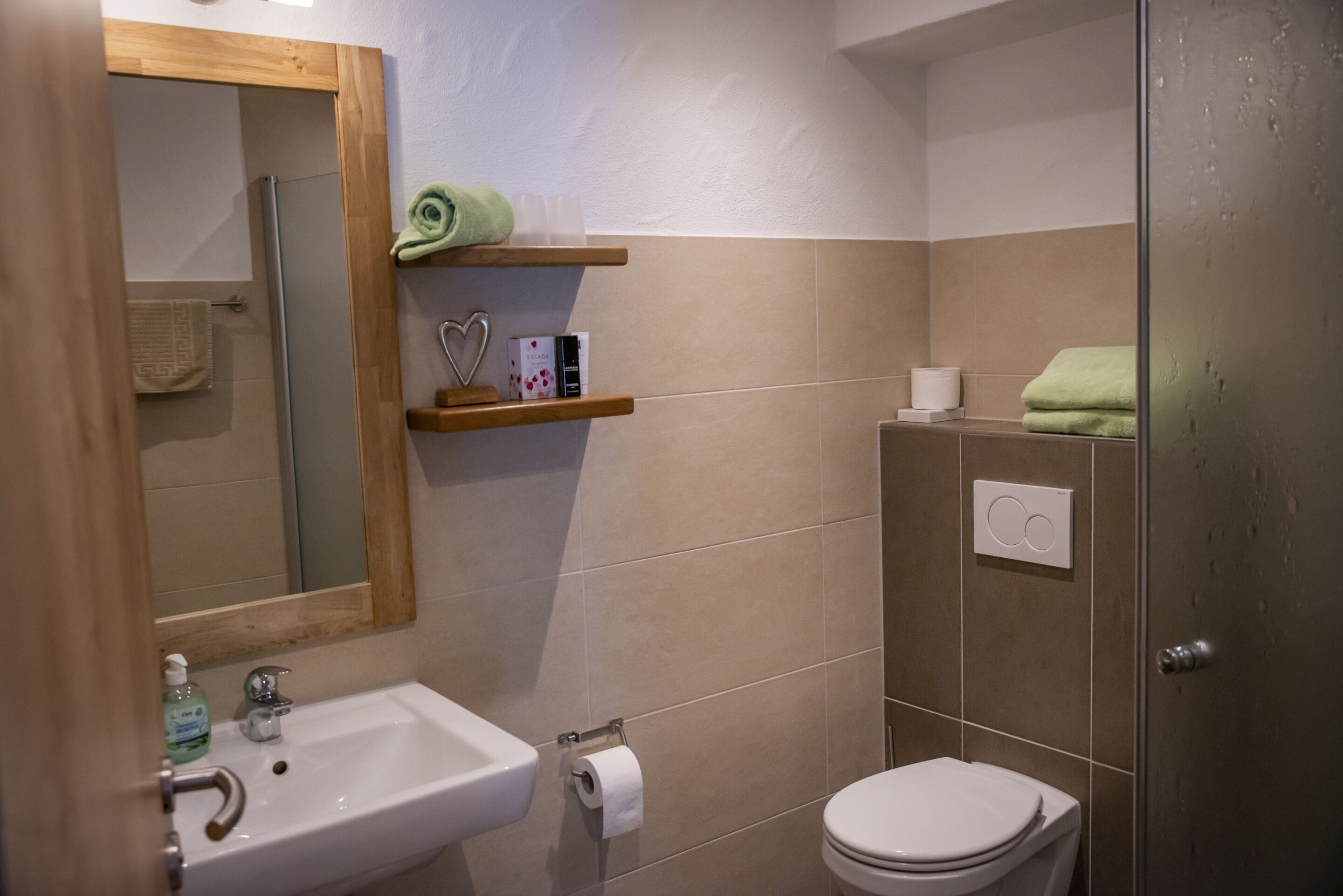 Ein Badezimmer mit Waschbecken, Toilette und Dusche. Handtücher und Pflegeprodukte liegen auf Regalen.