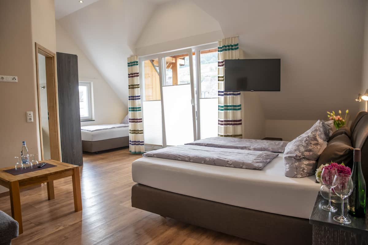 A bedroom with a bed, a TV mounted on the wall, a wooden table with a bottle and glasses, and a window with curtains.