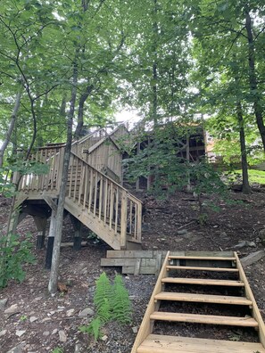 Eine Holztreppe führt zu einer Holzdiele mit Geländer, umgeben von Bäumen und einem Gebäude im Hintergrund.