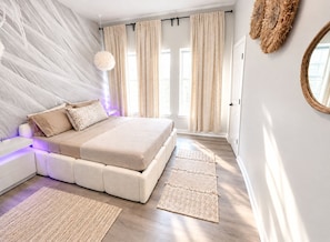 Une chambre à coucher comprenant un lit, des tables de chevet, un luminaire suspendu et une grande fenêtre avec des rideaux.