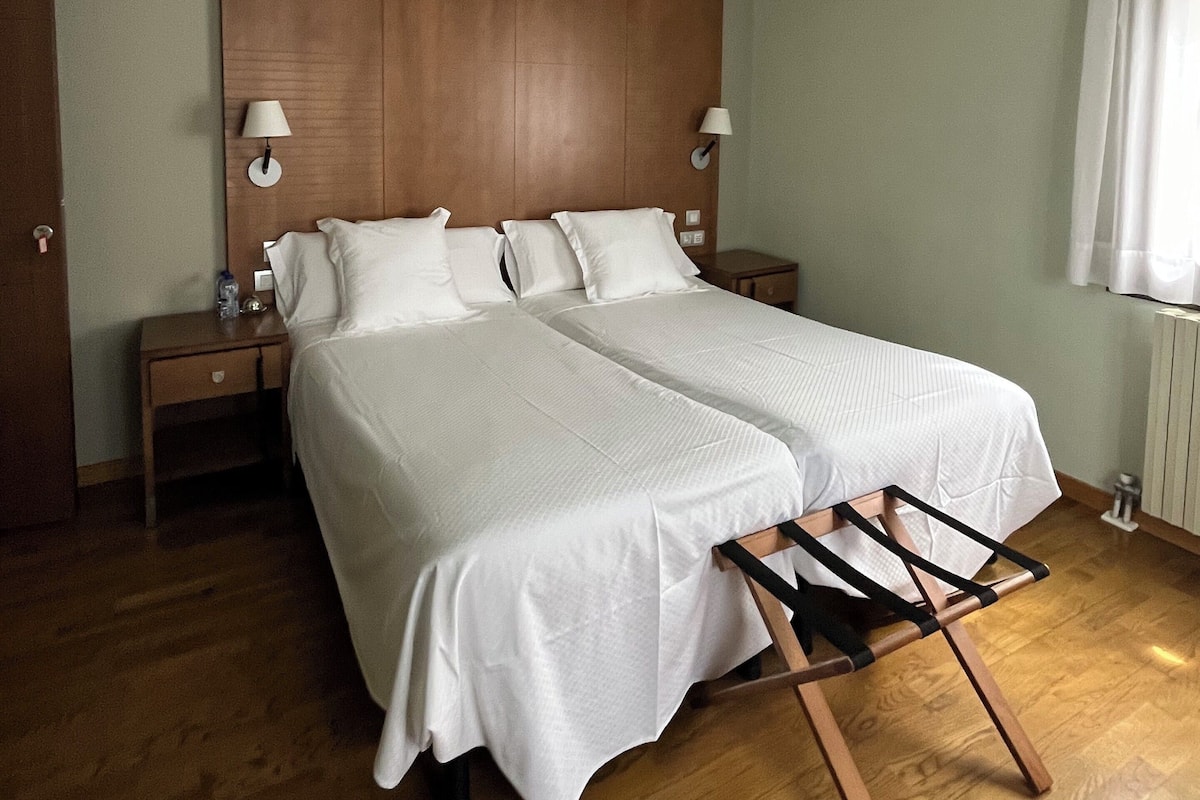 Cama de casal com roupa de cama branca, duas mesas de cabeceira com luminárias, cabeceira de madeira e piso de madeira.